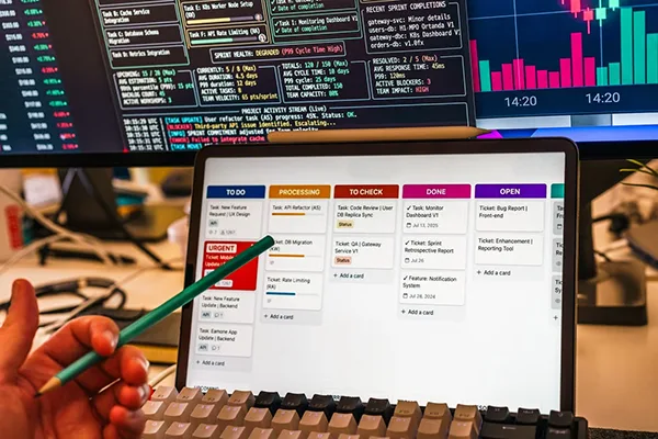 A hand holding a tablet whose screen shows a software interface with tasks lined up and a half computer screen with some statistics visible in the background.
