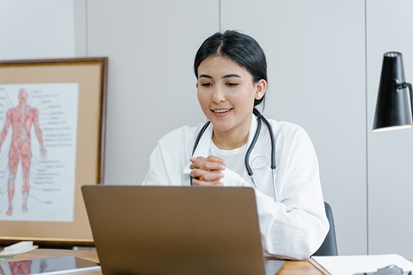 A smiling doctor sitting with crossed hands watching on the laptop placed open on the table in a clinic background